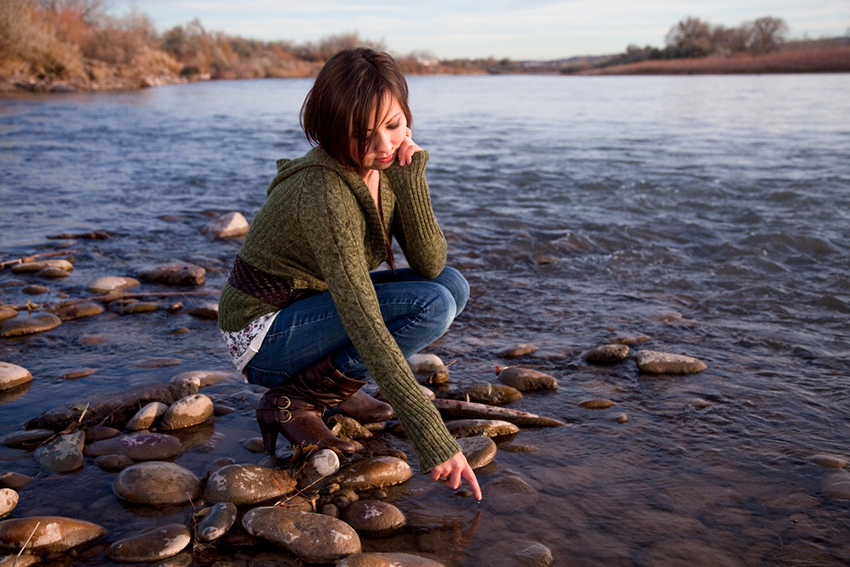 Cute teenage girl, crouching on river bank, dreamily lets a water droplet fall from her finger