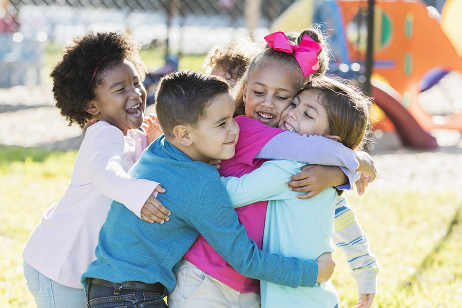 A multi-ethnic group of children playing outdoors on a playground on a sunny day. They are all playfully hugging each other.