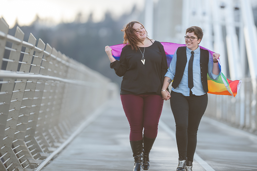 A young adult lesbian couple in their 20's are walking and smiling while draped in a pride flag